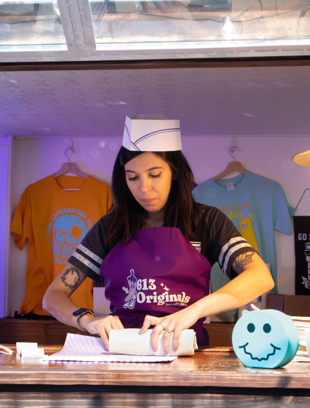 A woman rolling up a tote bag wearing a paper hat and a apron that says "613 Originals. Hot off the press"