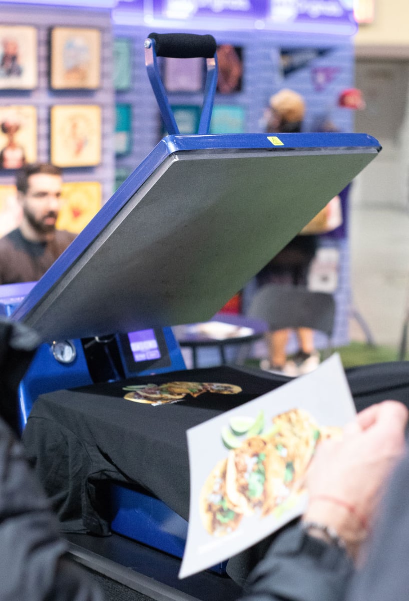 A person holding our Direct to Film taco transfers and looking at a blue heat press. A wall with different completed garments is seen in the background.