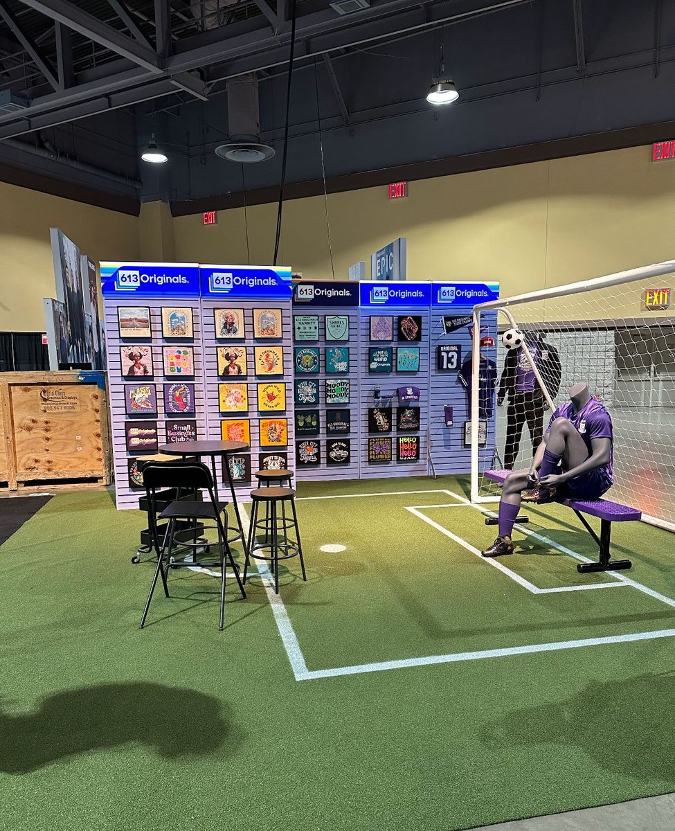 A fake turf with a soccer net. Inside the net is a soccer bench with a mannequin sitting on it. The mannequin is wearing a decorated uniform. At the far end is a palette wall with various transfer types displayed.