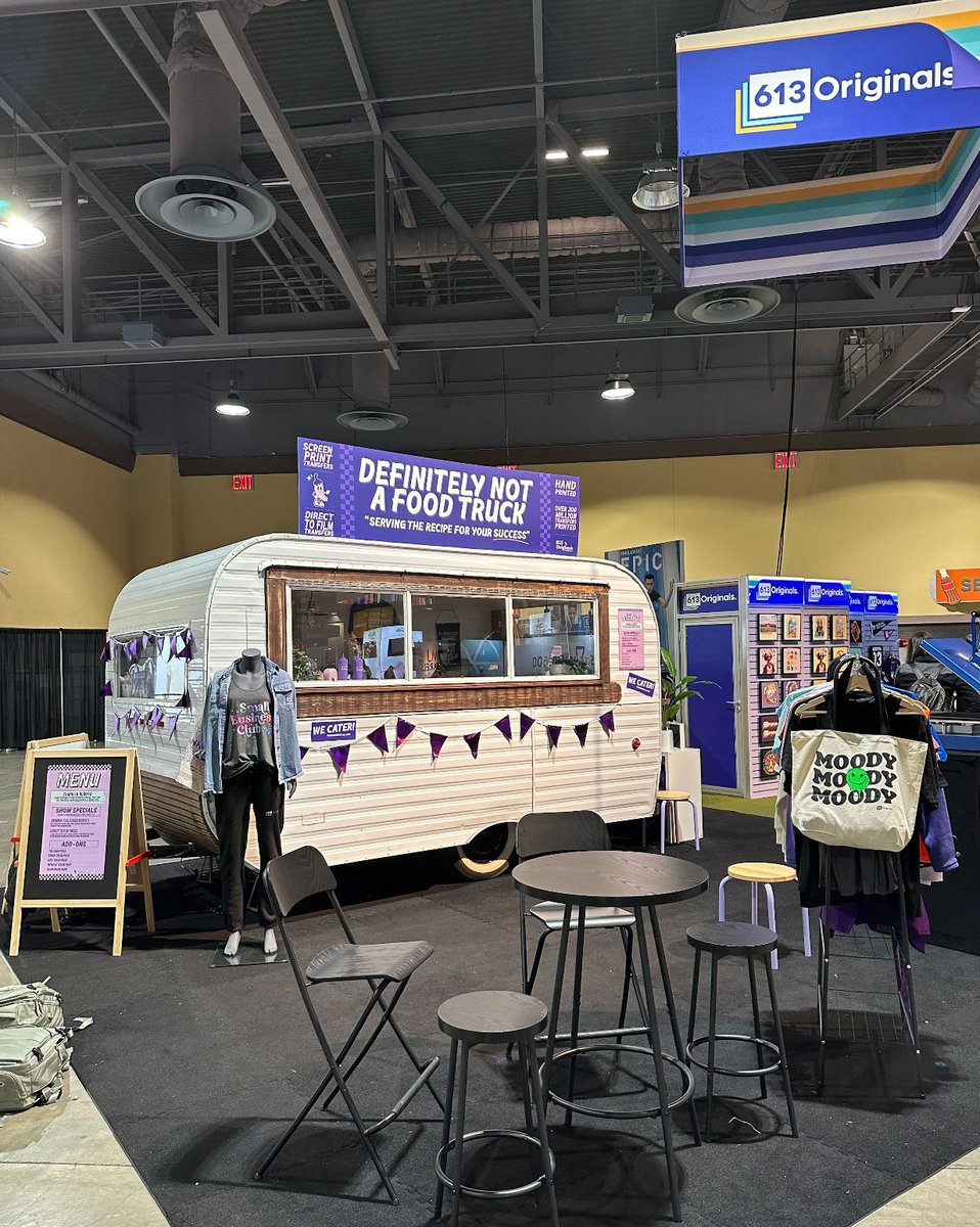 A food truck with a sign that says "Definitely not a food truck." is visible. A table and chairs are in front with a garment rack full of finished, decorated apparel next to it.