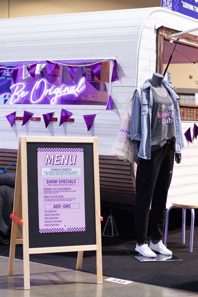 A neon sign reads "613 Originals" and a menu board is visible with various options. A mannequin is standing next to the menu board wearing various decorated apparel.