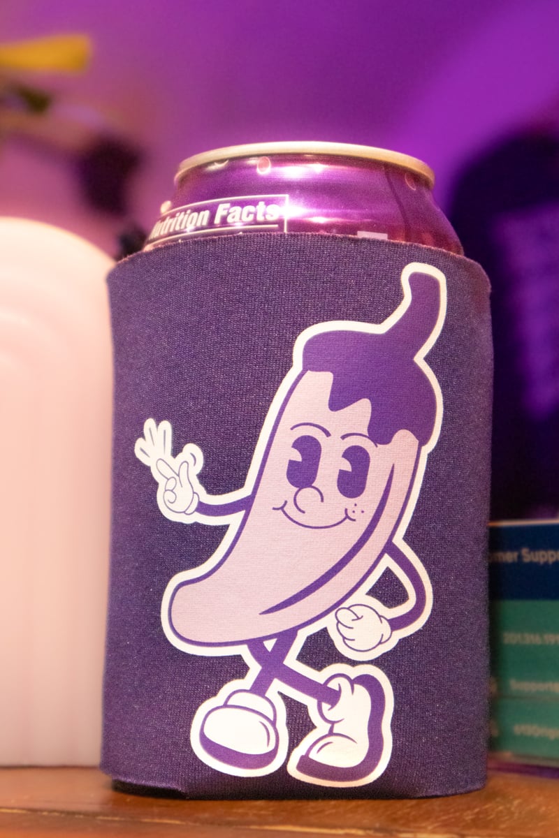 A purple can cooler holder with a mascot in the shape of a pepper snapping his fingers.
